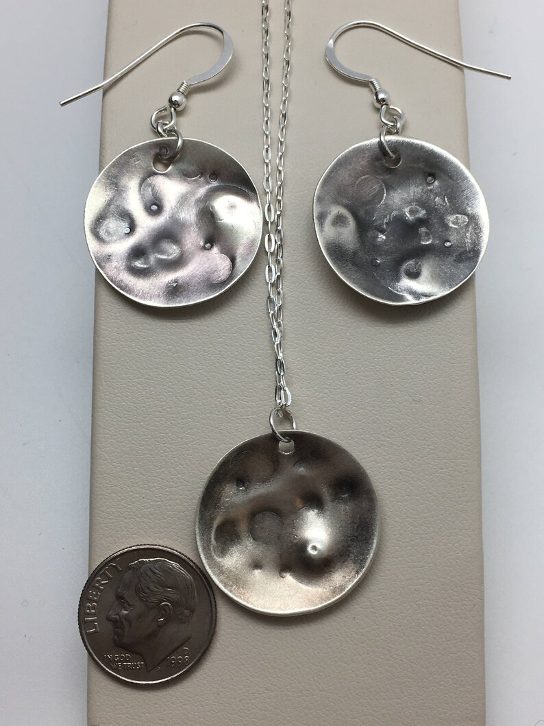 May include: A silver necklace and earrings set with round, textured pendants. The pendants are about the size of a US dime.