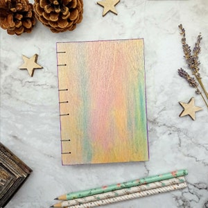 May include: A rainbow-coloured wooden journal with a purple edge and a stitched binding. The journal is lying on a white marble surface with wooden stars and a sprig of lavender.