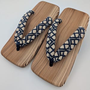 May include: A pair of traditional Japanese geta sandals. The wooden platforms have a natural wood grain finish. The straps are patterned with a navy blue and cream-colored geometric design.