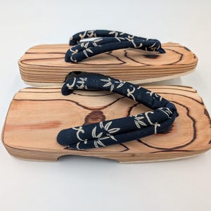 May include: Pair of wooden geta sandals. The sandals have a light brown wood base with a dark blue fabric strap featuring a white floral pattern. The sandals are designed with a traditional Japanese style.