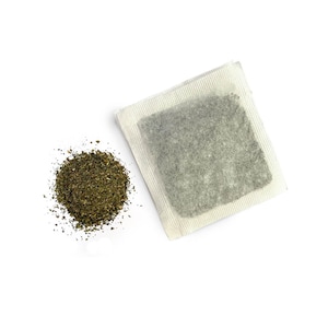 May include: A white tea bag with loose green tea leaves scattered around it on a white background.