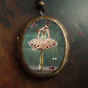 May include: A vintage-style locket pendant with a detailed brass frame. Inside the locket is a colorful illustration of a ballerina in a white and red dress, holding a red ribbon and balancing on a small red stool.