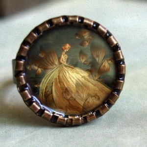May include: A round ring with a bronze-colored metal band. The ring's face features a vintage-style image of a woman in a golden gown. The image is set against a dark background with floating golden elements.