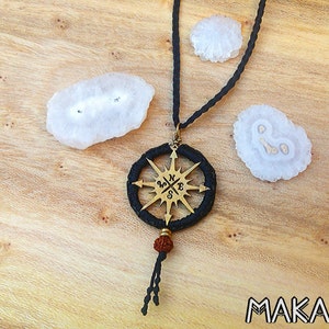 May include: A brass compass pendant necklace with a black cord and a brown bead. The compass has a black background with gold accents and the letters N, S, E, and W. The necklace is surrounded by three white quartz crystals.