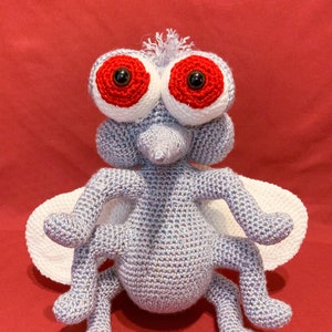 May include: A crocheted plush toy fly with large red eyes and white wings. The fly is sitting on a red surface.