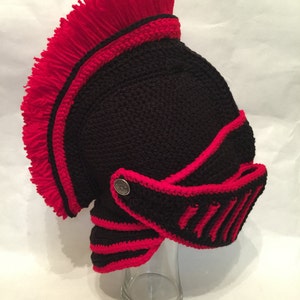 Peut inclure: Un casque en crochet noir et rouge avec une frange rouge sur le dessus. Le casque a une visière et un bouton métallique sur le côté.