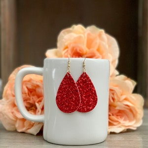 May include: A pair of red glitter teardrop earrings hanging from a white mug. The earrings are made of red glitter and have a teardrop shape. The mug is white and has a handle.