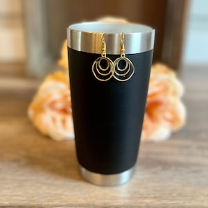 May include: Gold-toned earrings with a triple circle design. The earrings are hanging from a black tumbler.
