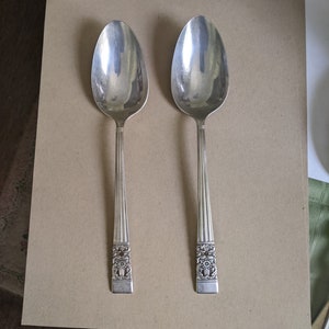 Puede incluir: Dos cucharas de plata con mangos ornamentados. Las cucharas están sobre una superficie marrón.