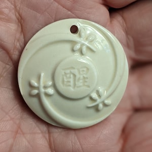 May include: A circular off-white pendant featuring a central embossed circle with two Chinese characters. The surface is decorated with three raised dragonfly motifs arranged in a swirling pattern. A small hole is positioned at the top for hanging.