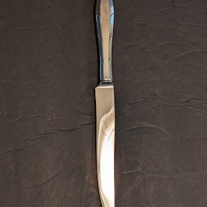 May include: A stainless steel butter knife with a decorative handle. The handle has a rounded, elongated shape with a central groove and vertical lines. The blade is smooth and curved.