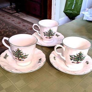 Puede incluir: Tres tazas de té blancas y platillos con un diseño de árbol de Navidad. Las tazas tienen un asa y se asientan sobre platillos a juego con un borde verde. El árbol de Navidad está decorado con adornos y regalos coloridos.