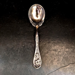 Könnte beinhalten: Ein silberner Löffel mit einer abgerundeten Schale und einem detaillierten Blumendesign am Griff. Der Löffel hat eine reflektierende Oberfläche und ist wahrscheinlich aus Sterlingsilber oder einem ähnlichen Metall gefertigt. Der Griff weist geprägte Blumen auf.