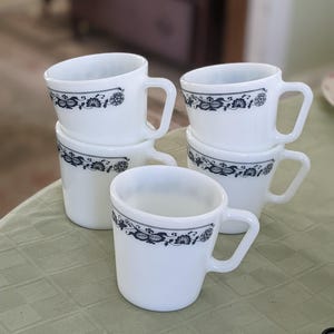 Könnte beinhalten: Ein Stapel von vier weißen Keramikbechern mit einem schwarzen Blumendesign. Die Becher haben eine abgerundete Form und einen Henkel auf der rechten Seite.