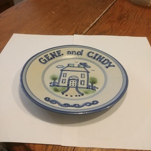 Puede incluir: Un plato de cerámica con un borde azul y blanco y una casa amarilla con molduras verdes y una puerta azul. El texto "GENE y CINDY" está escrito en azul alrededor de la casa.