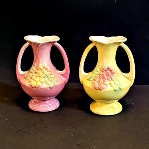 May include: Two ceramic vases, one pink and one yellow, each with a floral design and two handles. The vases have a pedestal base and a ruffled top. The pink vase has yellow flowers, and the yellow vase has pink flowers.