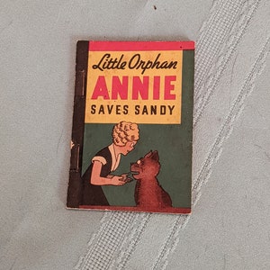 Pode incluir: Uma capa de livro vintage de Little Orphan Annie com um esquema de cores vermelho, amarelo e verde. O t&iacute;tulo diz "Little Orphan Annie Saves Sandy". A capa apresenta uma ilustra&ccedil;&atilde;o de Annie, uma menina com cabelo loiro, e seu cachorro Sandy.