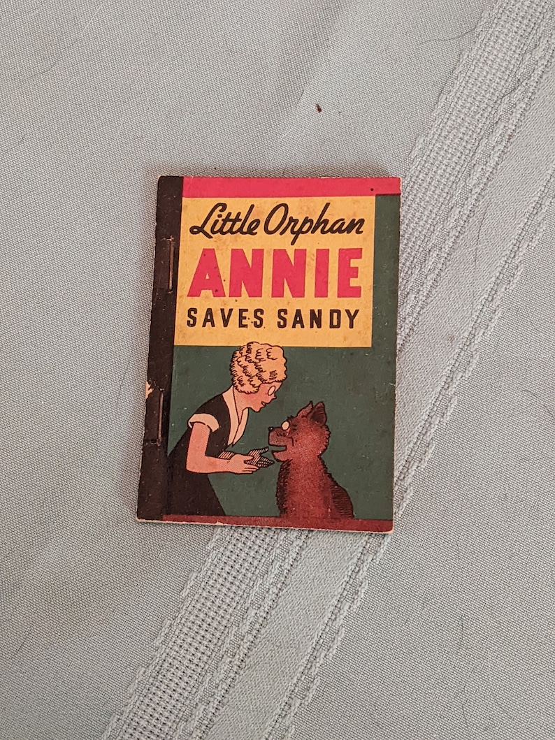 Pode incluir: Uma capa de livro vintage de Little Orphan Annie com um esquema de cores vermelho, amarelo e verde. O t&iacute;tulo diz "Little Orphan Annie Saves Sandy". A capa apresenta uma ilustra&ccedil;&atilde;o de Annie, uma menina com cabelo loiro, e seu cachorro Sandy.