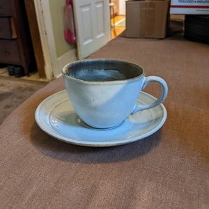 Puede incluir: Una taza de café de cerámica blanca con un borde marrón y un platillo a juego. La taza está llena de café y está sobre una mesa marrón.