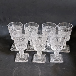 Puede incluir: Un juego de ocho copas de agua transparentes de estilo vintage. Cada copa presenta una base cuadrada, un tallo detallado y un cuenco estampado. Las copas están dispuestas sobre una superficie oscura, mostrando su intrincado diseño.