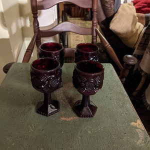 Puede incluir: Cuatro copas de vino de vidrio rojo oscuro con un diseño texturizado se encuentran sobre una mesa de madera verde. Las copas tienen una copa redonda y un tallo alto y delgado.