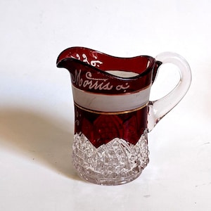 Puede incluir: Jarra de vidrio vintage con asa transparente y borde y base rojo rubí. La jarra tiene una banda blanca con el nombre "Morris" en cursiva. La base tiene un patrón de corte de diamante.