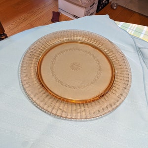 May include: A clear glass serving plate with a raised, textured rim and a delicate etched design of leaves and a flower in the center. The plate is amber in color.