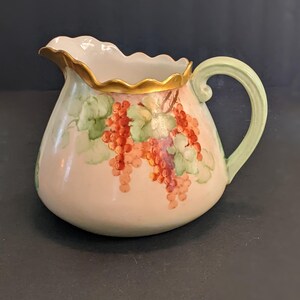 May include: A vintage ceramic pitcher with a light pink base, featuring hand-painted red grapes and green leaves. The rim is scalloped and trimmed with gold, and the handle is light green. A decorative piece for serving.