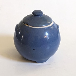 以下が含まれることがあります： 小さな青いセラミックジャーとそれに合う蓋。ジャーは丸みを帯びたボディで、小さなハンドルが2つと白い縁があります。蓋には小さなノブが付いています。ジャーは無地のマットブルーです。