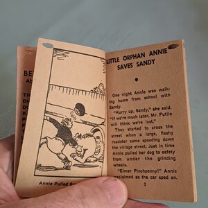 Pode incluir: Uma ilustra&ccedil;&atilde;o em preto e branco de uma menina puxando um c&atilde;o para fora do caminho de um carro. O texto "Little Orphan Annie Saves Sandy" est&aacute; no topo da p&aacute;gina. O texto "Annie Pulled S- " est&aacute; na parte inferior da p&aacute;gina.