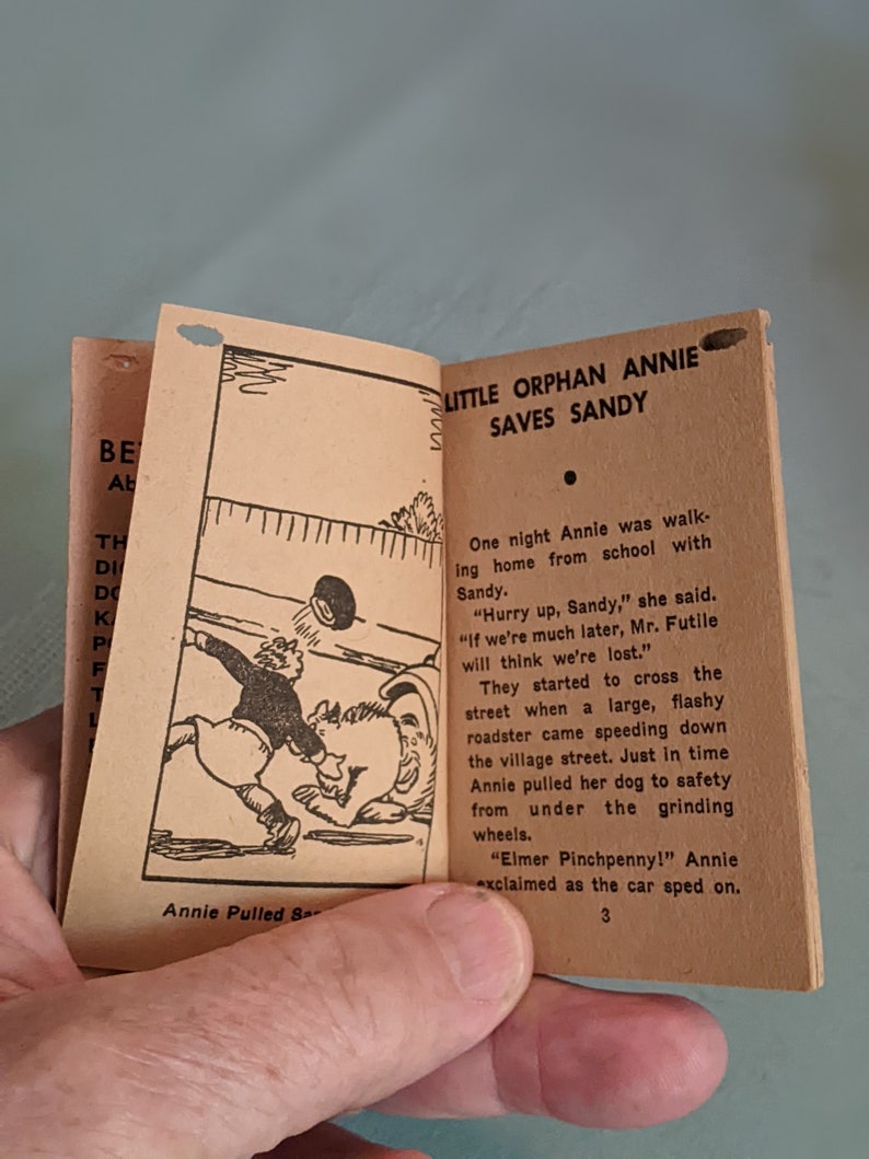 Pode incluir: Uma ilustra&ccedil;&atilde;o em preto e branco de uma menina puxando um c&atilde;o para fora do caminho de um carro. O texto "Little Orphan Annie Saves Sandy" est&aacute; no topo da p&aacute;gina. O texto "Annie Pulled S- " est&aacute; na parte inferior da p&aacute;gina.