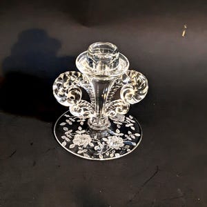 Peut inclure: Bougeoir en verre transparent avec un motif floral décoratif sur la base. Le bougeoir présente un design évasé et orné avec une base circulaire et une coupelle pour tenir une bougie. La base présente des motifs floraux et de feuilles gravés.