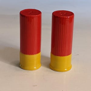 May include: A pair of red and yellow salt and pepper shakers. The shakers are cylindrical, with a red top and a yellow base. Each shaker has two small holes on the top.