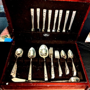 Pode incluir: Um conjunto de talheres em tom prateado disposto numa caixa de armazenamento de madeira castanho-escura. O conjunto inclui várias colheres, garfos e facas com cabos decorativos. O interior da caixa é forrado com tecido castanho-escuro.
