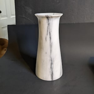 May include: A tall, white marble vase with black veining. The vase has a flared top and a gently tapered body. The polished surface reflects light, highlighting the natural patterns of the stone. This decorative vase is suitable for displaying flowers or as a standalone accent piece.