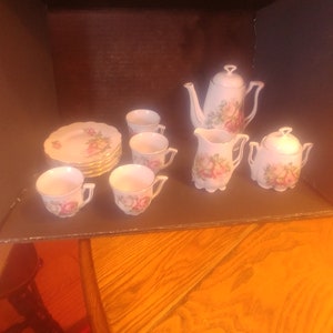 Puede incluir: Un juego de té de porcelana blanca con un diseño floral. El juego incluye una tetera, una jarra de crema, un azucarero, cuatro tazas y cuatro platillos.