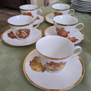 May include: Set of six white teacups and saucers with gold trim and fruit illustrations. The cups and saucers feature a variety of fruits, including apples, pears, and grapes.