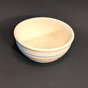 Puede incluir: Un cuenco pequeño de cerámica blanquecina con forma redondeada. El cuenco presenta dos rayas horizontales, una azul claro y otra rosa, que rodean el exterior. El interior del cuenco es de color blanquecino liso.