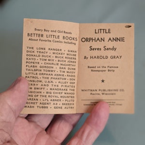 Pode incluir: Uma capa de livro de bolso vintage para "A Pequena &Oacute;rf&atilde; Annie Salva Sandy" de Harold Gray. A capa &eacute; marrom com texto branco e uma estrela. O livro &eacute; baseado na famosa tira de jornal.