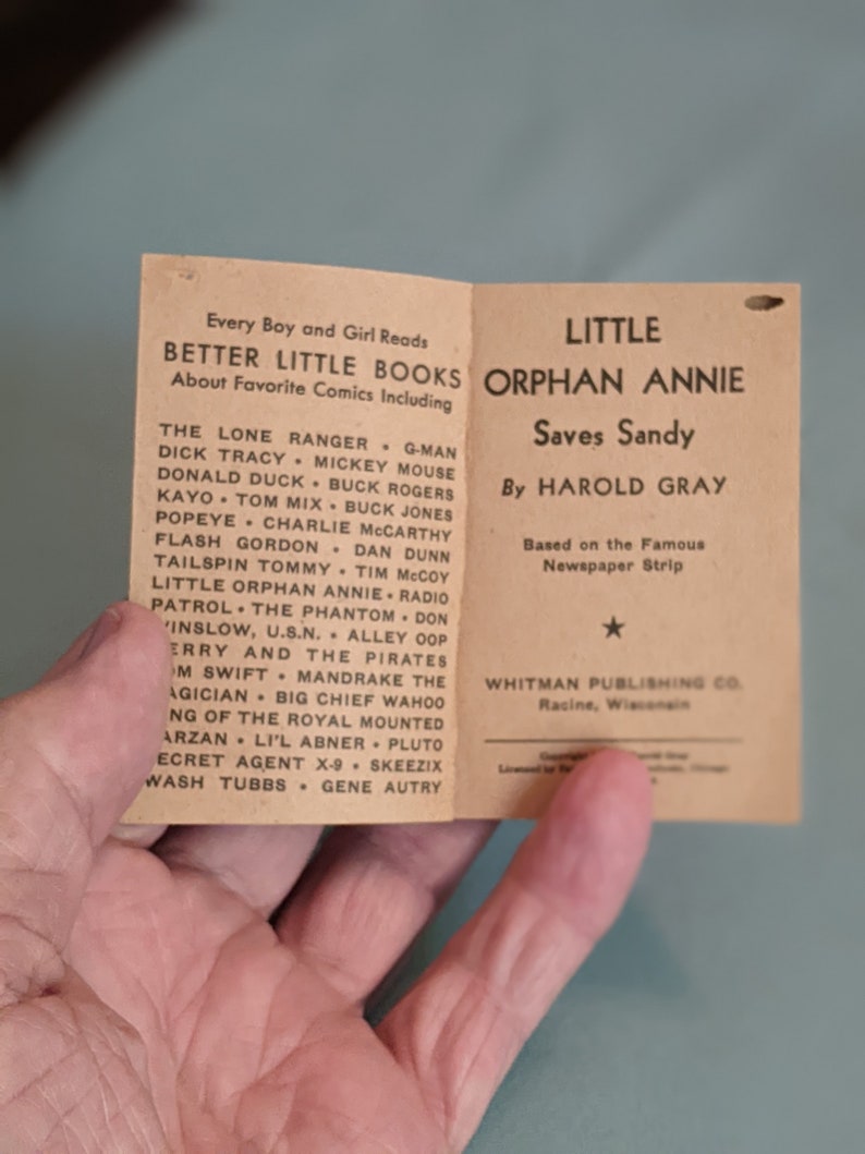 Pode incluir: Uma capa de livro de bolso vintage para "A Pequena &Oacute;rf&atilde; Annie Salva Sandy" de Harold Gray. A capa &eacute; marrom com texto branco e uma estrela. O livro &eacute; baseado na famosa tira de jornal.