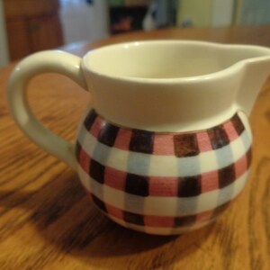May include: A white ceramic milk jug with a pink, blue and brown checked pattern. The jug has a rounded body and a handle.