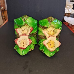 Puede incluir: Dos sujetalibros florales de cerámica. Cada uno presenta una base de hojas verdes, una flor color crema con bordes rojos y un tallo marrón. Los sujetalibros están diseñados para parecerse a un arreglo floral estilizado.