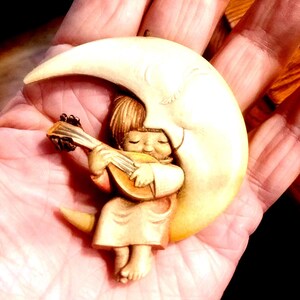 May include: A carved figurine of a child playing a lute while seated on a crescent moon. The figurine is in shades of beige and brown, with the child's face serene and eyes closed. The moon has a textured surface.