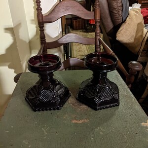 May include: Two dark red glass candle holders with a decorative pattern on the base. The candle holders are sitting on a green wooden table.