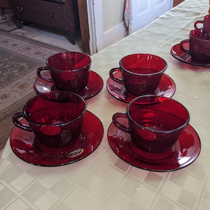 Puede incluir: Juego de seis tazas de té y platillos de vidrio rojo. Las tazas y los platillos son todos del mismo tamaño y forma. Están dispuestos en una fila sobre un mantel blanco.