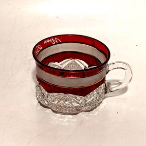 May include: A small glass cup featuring a deep red band around the upper rim, a clear glass handle, and a faceted diamond-cut pattern on the lower section. The red band includes a thin gold-colored horizontal line and white cursive text.