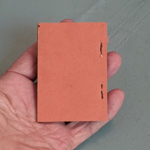 Pode incluir: Um pequeno livro retangular de cor laranja com encaderna&ccedil;&atilde;o costurada.