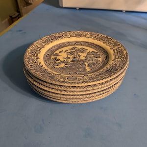 May include: A stack of five ceramic plates with a blue and white Willow pattern design. Each plate features a central landscape scene with a pagoda, bridge, and trees, surrounded by a decorative border. The plates are stacked on a blue surface.