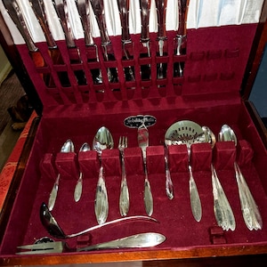 Op de afbeelding: Een vintage zilveren bestekset in een houten kist met een rode fluwelen binnenkant. De set bevat messen, vorken, lepels en serveerbestek. Het bestek heeft een glanzende, zilveren afwerking. De kist is open en toont de netjes gerangschikte stukken.