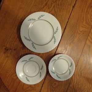 May include: Three white ceramic plates with a green and gold leaf design. The plates are nested together, with the largest plate on the bottom and the smallest plate on top.